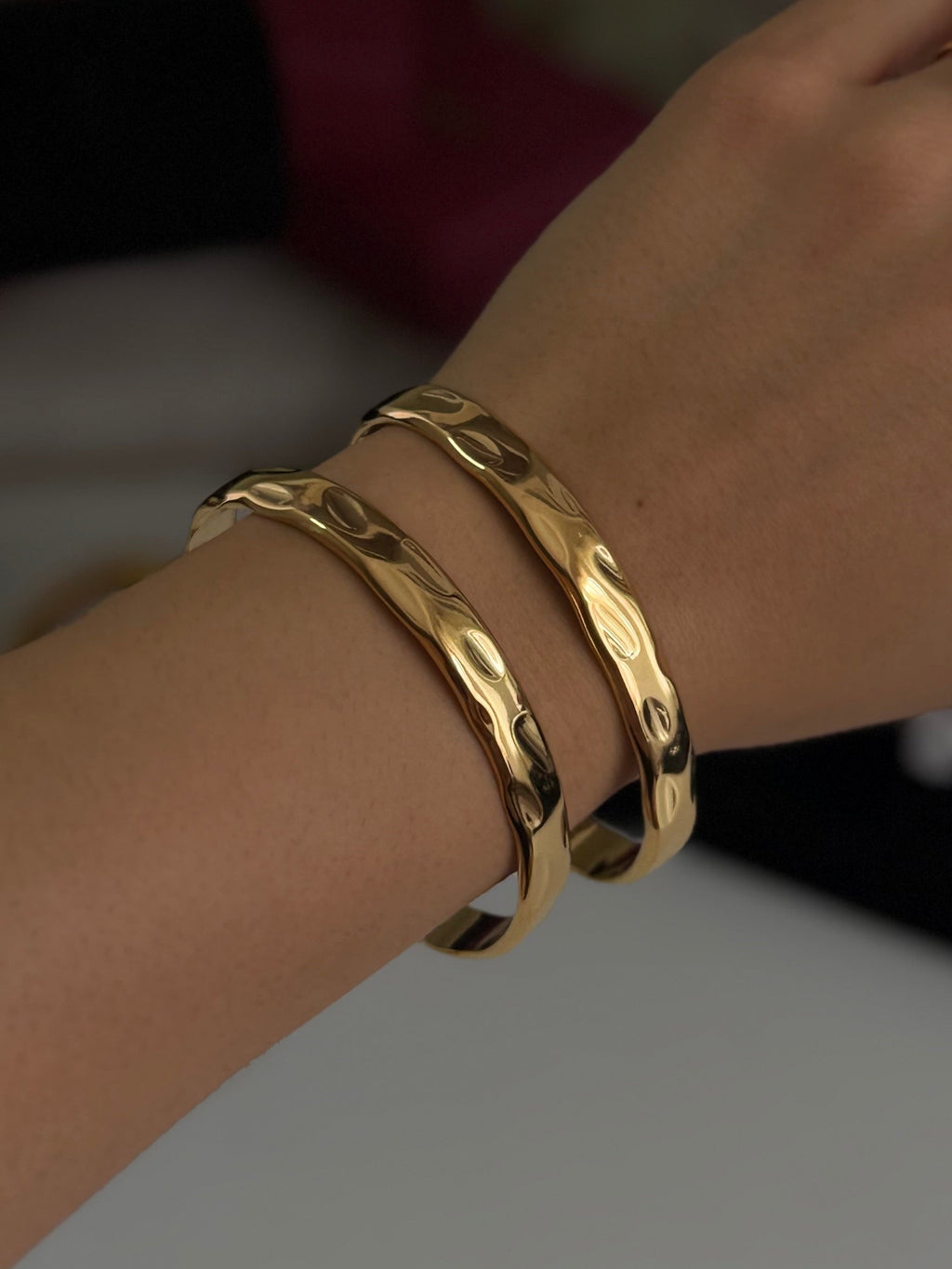 A close-up of a person's hand and wrist wearing two polished gold bangle bracelets, with two rings on the fingers, against a softly blurred indoor background.