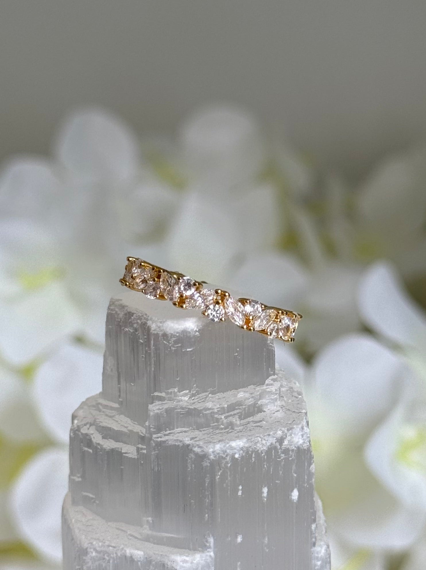 Gold ring with clear gemstone on a white crystal stick against a blurred floral background