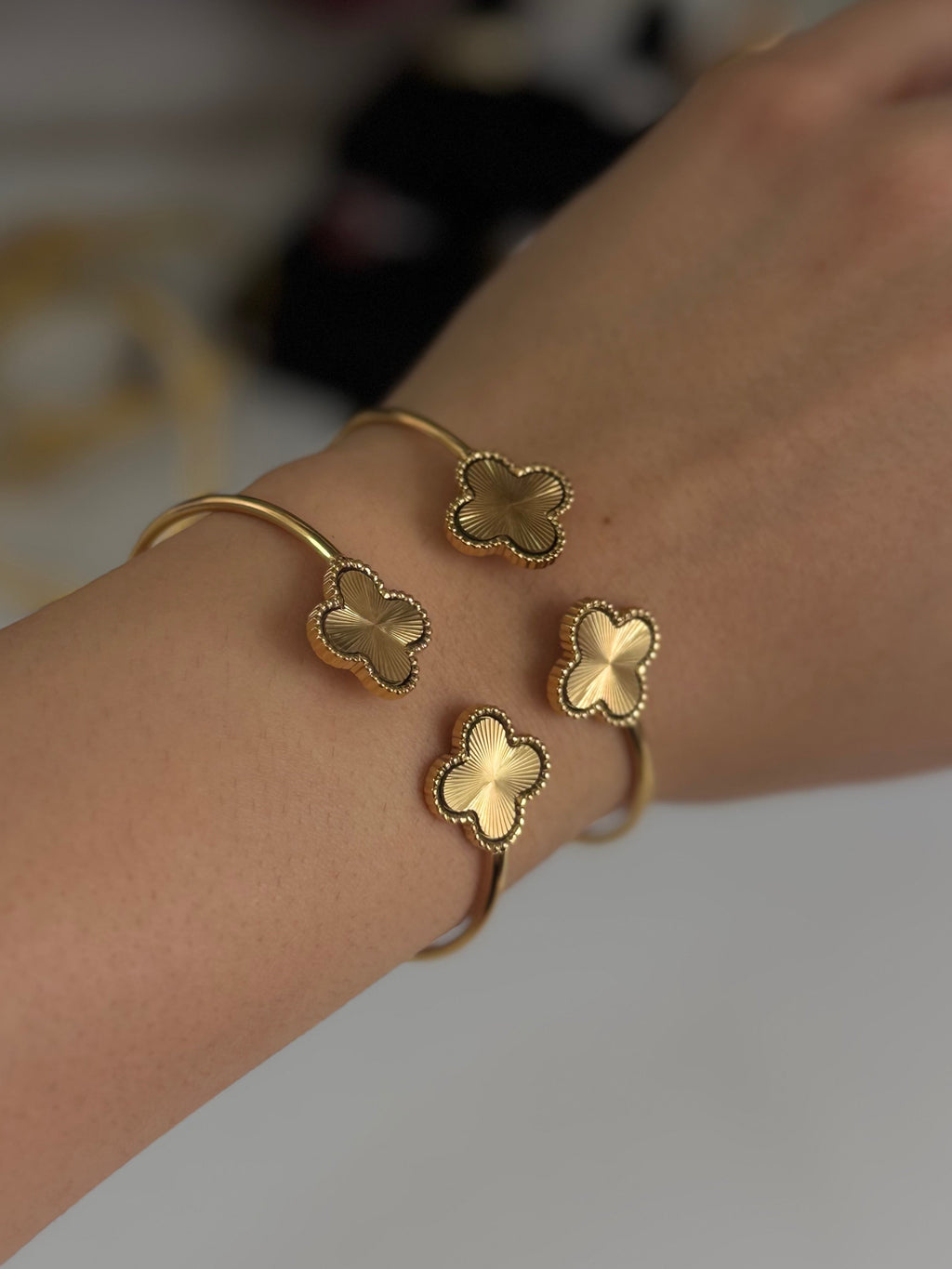 A close-up of a person's wrist wearing two gold-toned cuff bracelets, each adorned with three small clover-shaped charms.