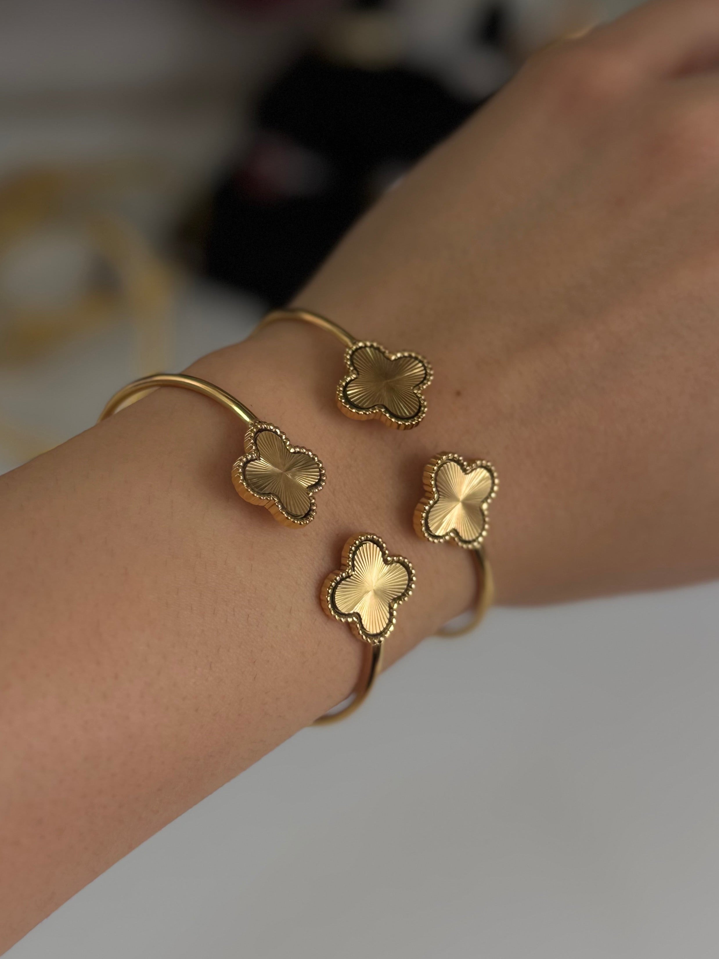 A close-up of a person's wrist wearing two gold-toned cuff bracelets, each adorned with three small clover-shaped charms.