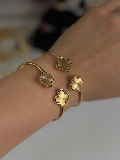 A close-up of a person's wrist wearing two gold-toned cuff bracelets, each adorned with three small clover-shaped charms.