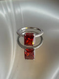 A silver ring featuring a large rectangular red gemstone and a smaller matching stone in a halo setting, displayed on a reflective white surface.