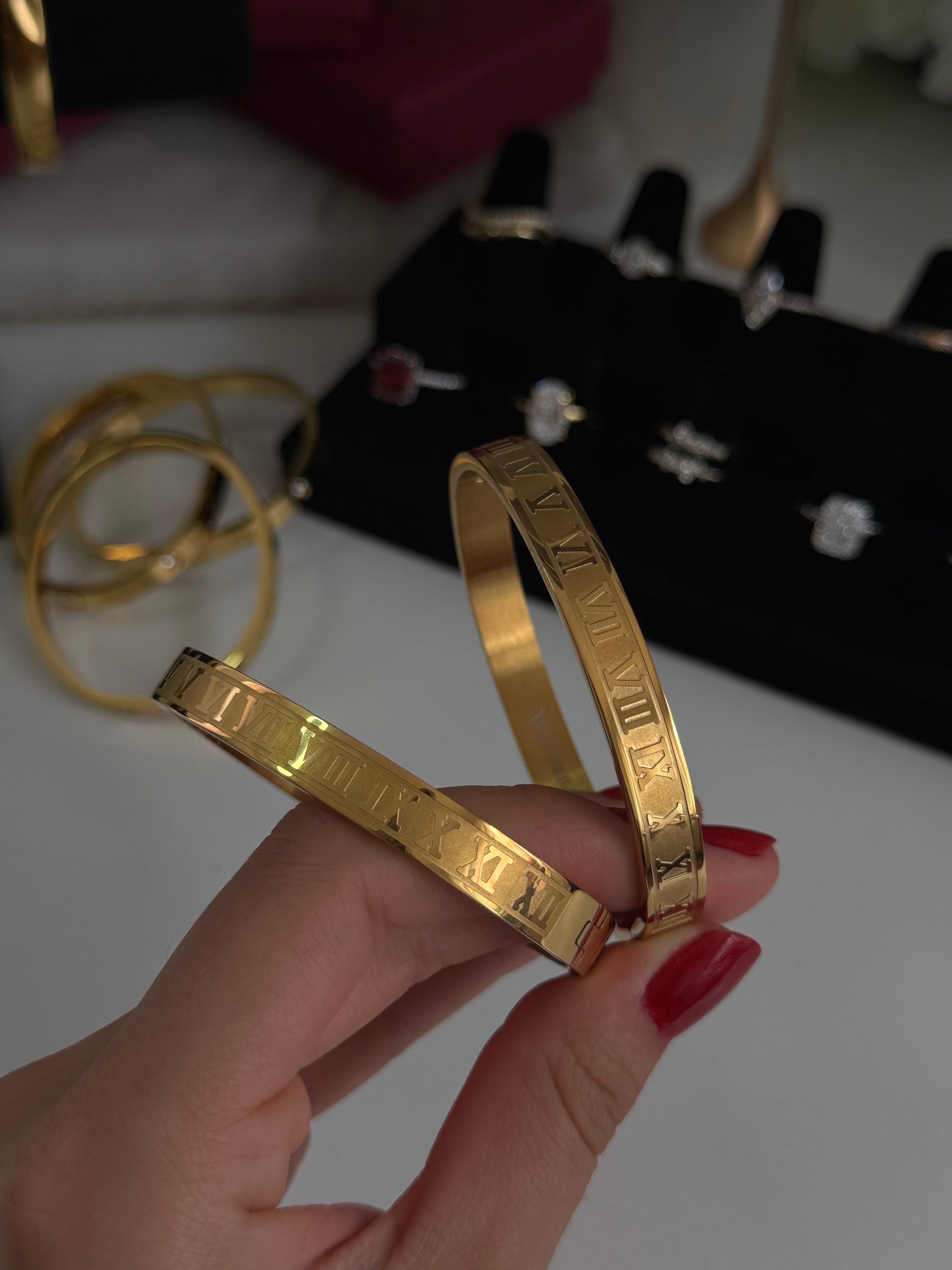 A hand with red nail polish holds two gold bangle bracelets with engraved Roman numerals, with a black jewelry display stand and other gold rings in the blurred background.