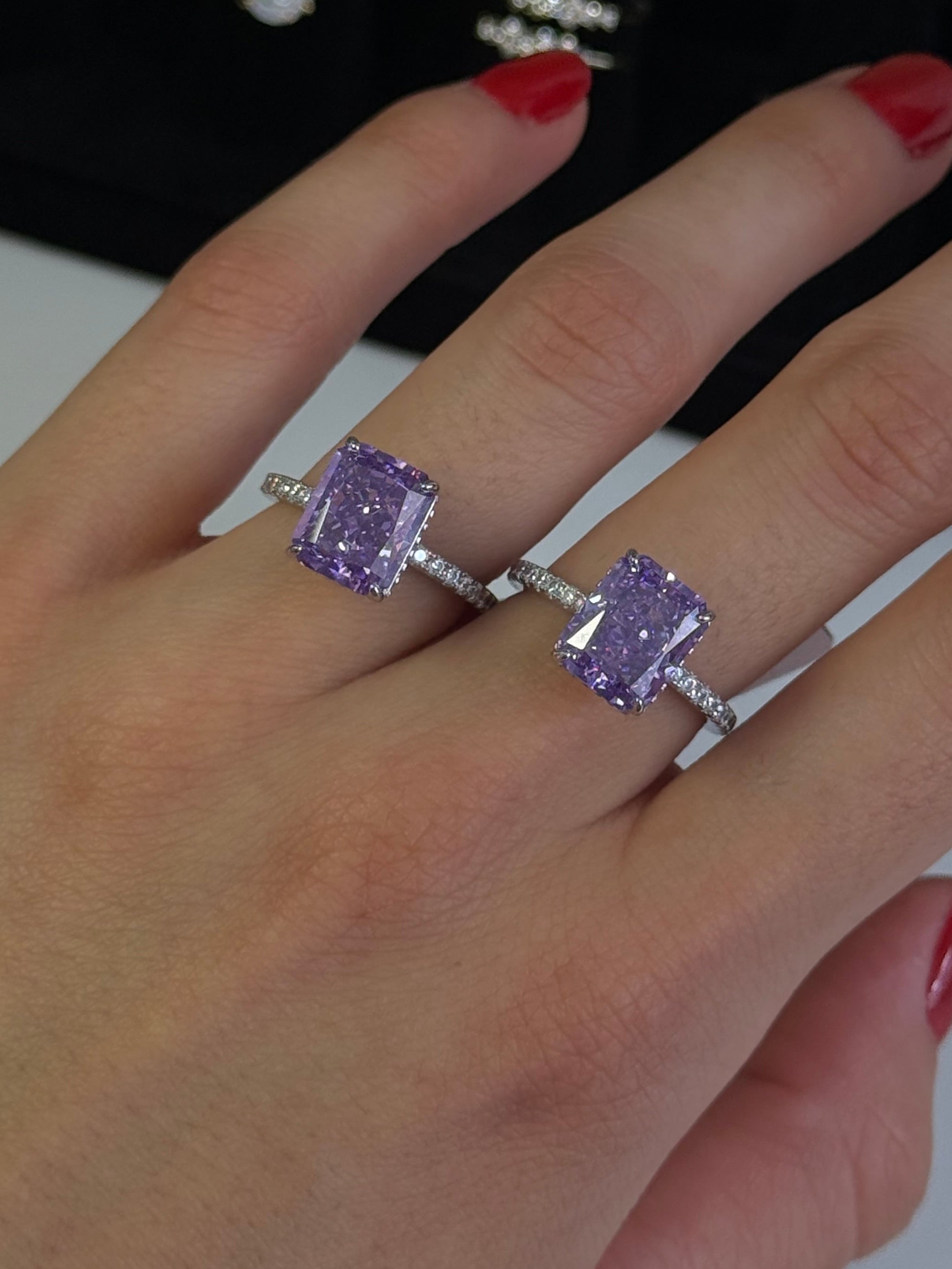 A close-up of a hand wearing a silver ring set with two rectangular light purple gemstones, with other jewelry displayed on a black stand in the background.