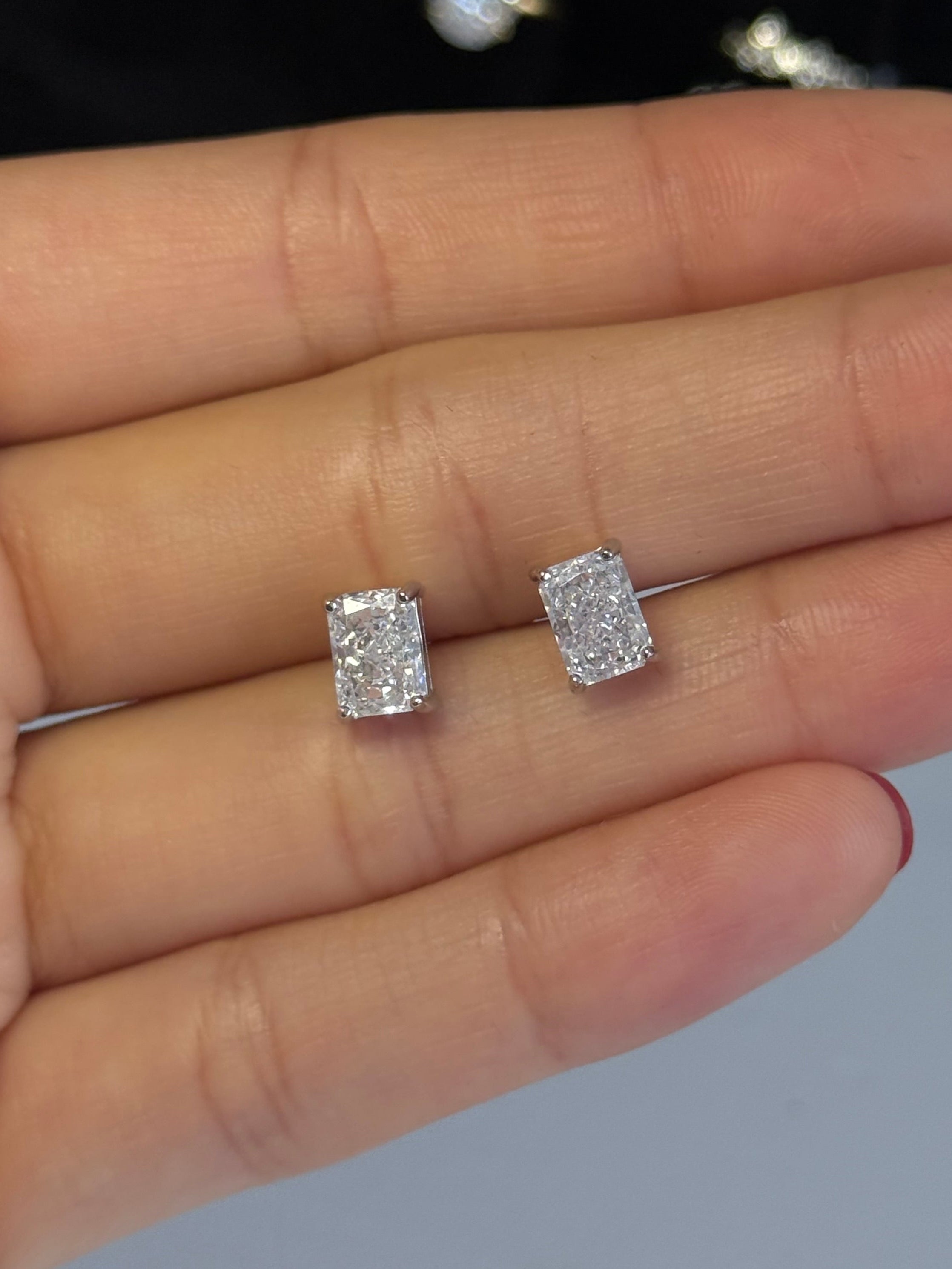 A close-up of a person's hand holding a pair of rectangular clear gemstone stud earrings in a silver-toned setting, with jewelry display pieces blurred in the background.