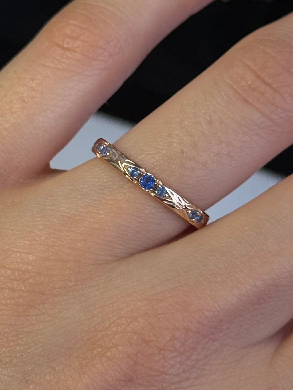 A close-up of a hand wearing a delicate silver band ring with small blue and yellow gemstones, with other jewelry displayed on a black stand in the blurred background.