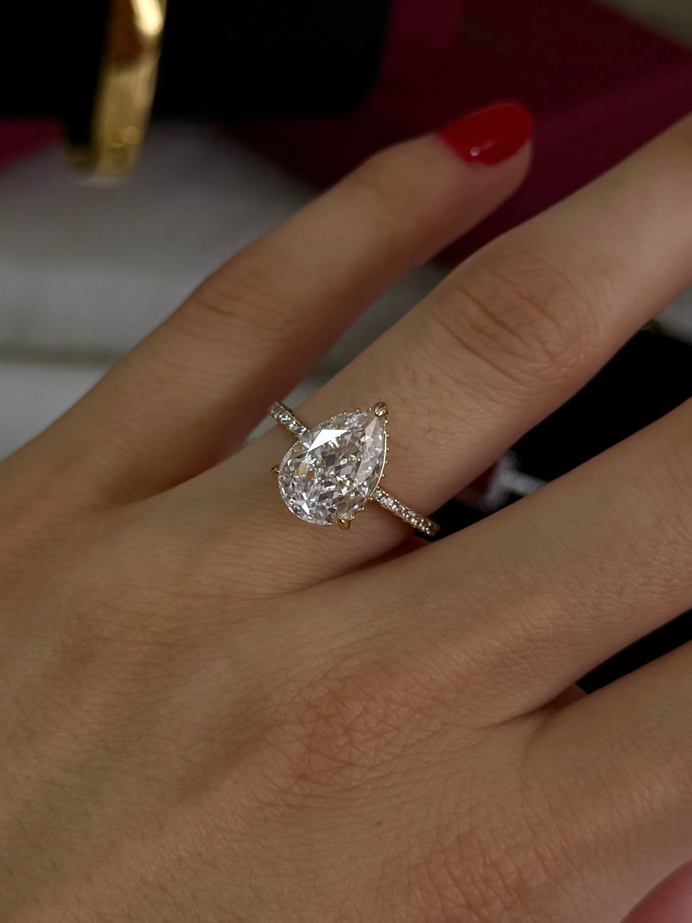 A close-up of a hand wearing a silver-toned ring with a pear-cut clear gemstone, with red nail polish and gold bracelets visible in the background.