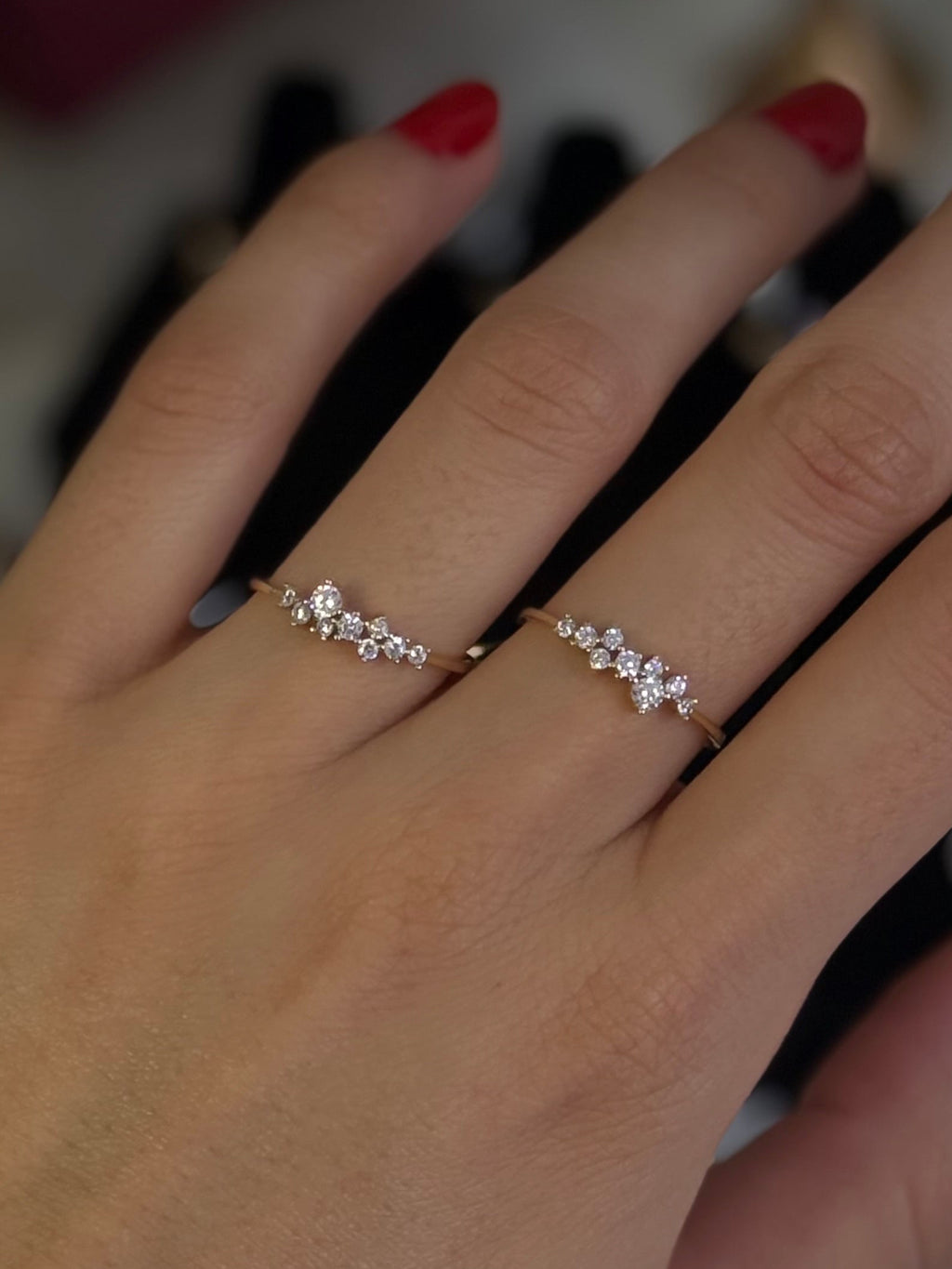 A close-up of a person's hand wearing two delicate silver rings with small clear stones and a gold bangle bracelet. The hand has red nail polish, and jewelry boxes are blurred in the background.