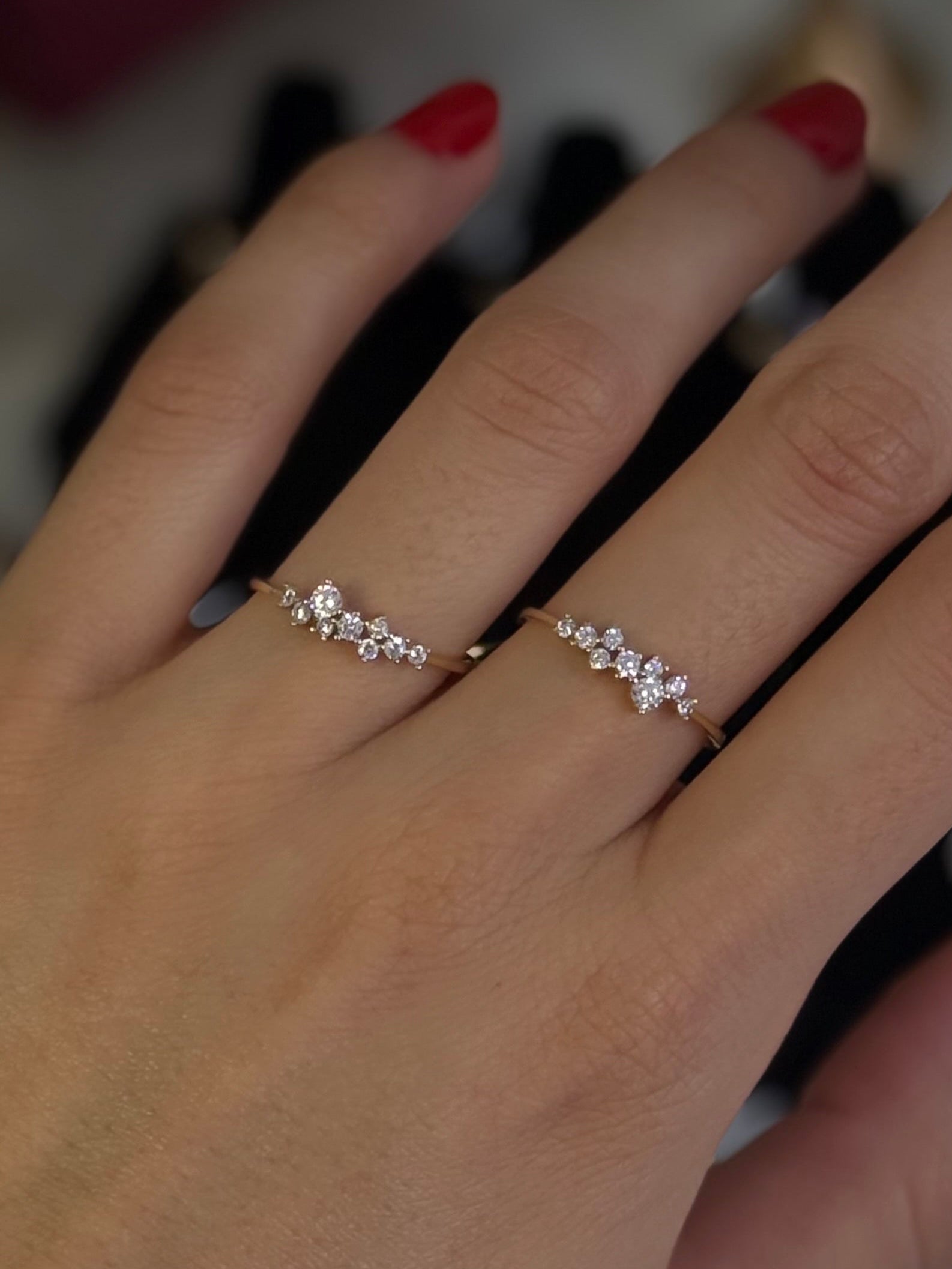 A close-up of a person's hand wearing two delicate silver rings with small clear stones and a gold bangle bracelet. The hand has red nail polish, and jewelry boxes are blurred in the background.