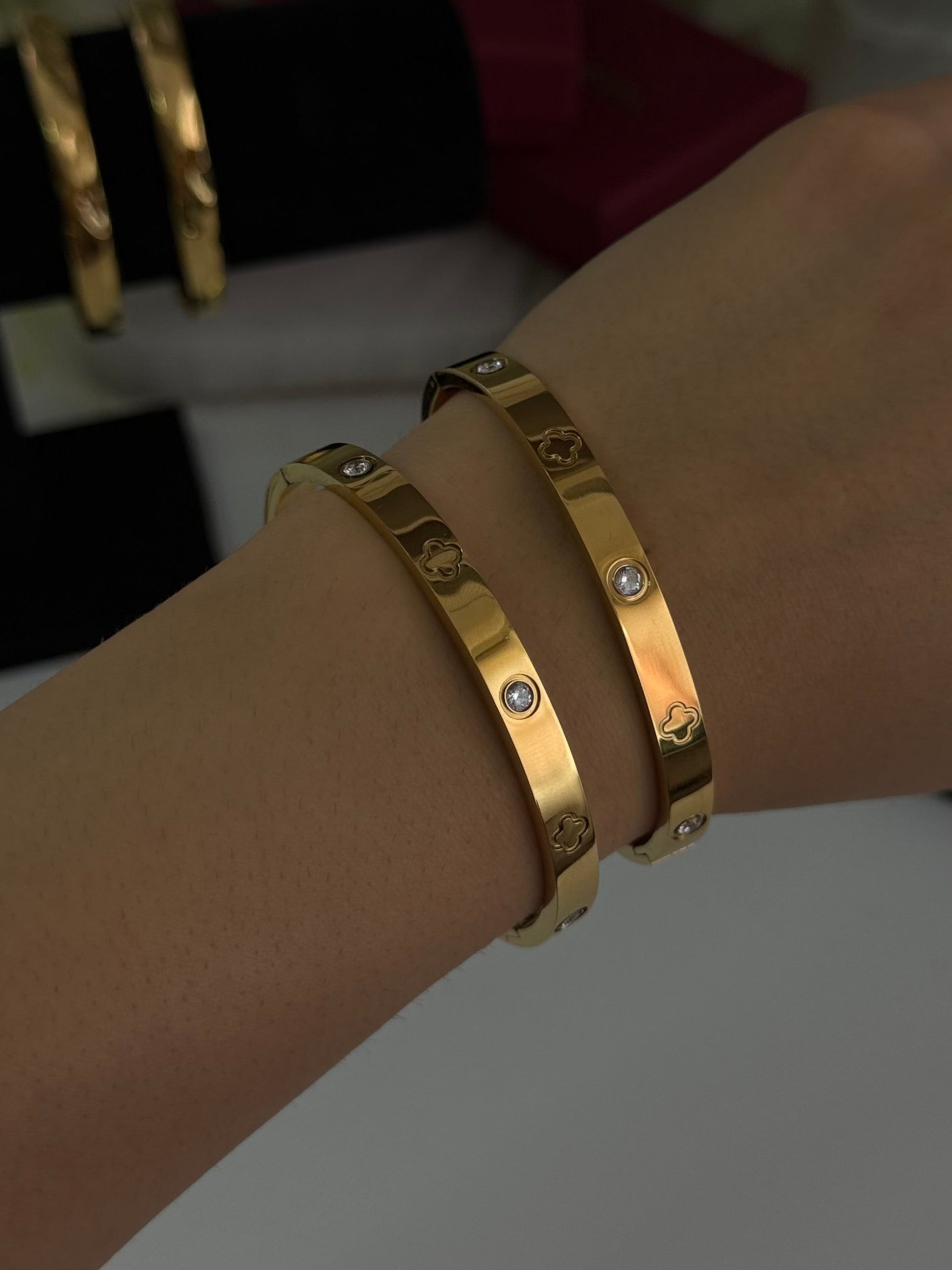 Two gold bangle bracelets with small clear stones, worn on a person's wrist, with a jewelry display stand and blurred background accessories.