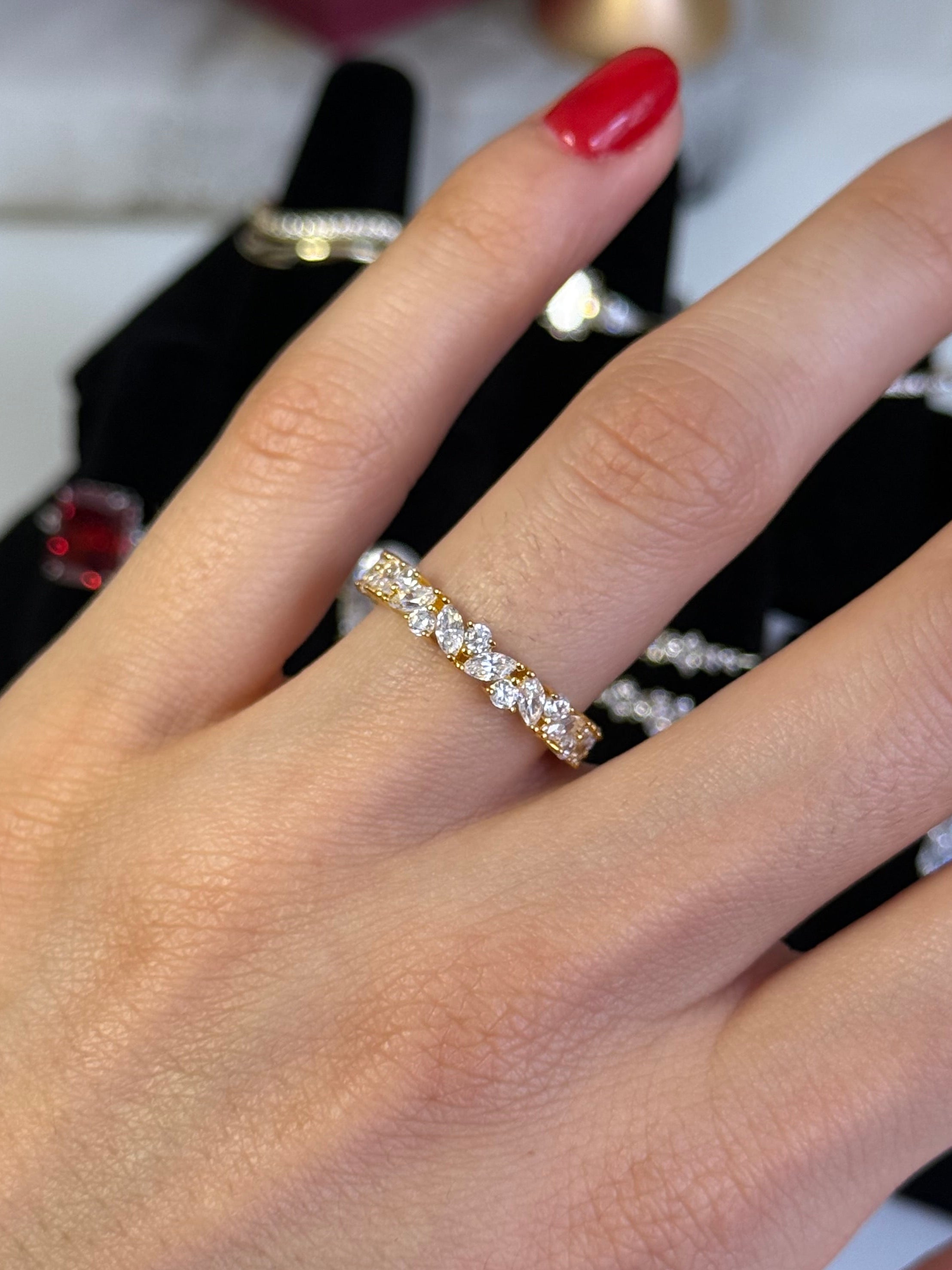 A close-up of a hand wearing a delicate gold eternity ring set with small clear stones, with red painted fingernails and jewelry displays in the background.