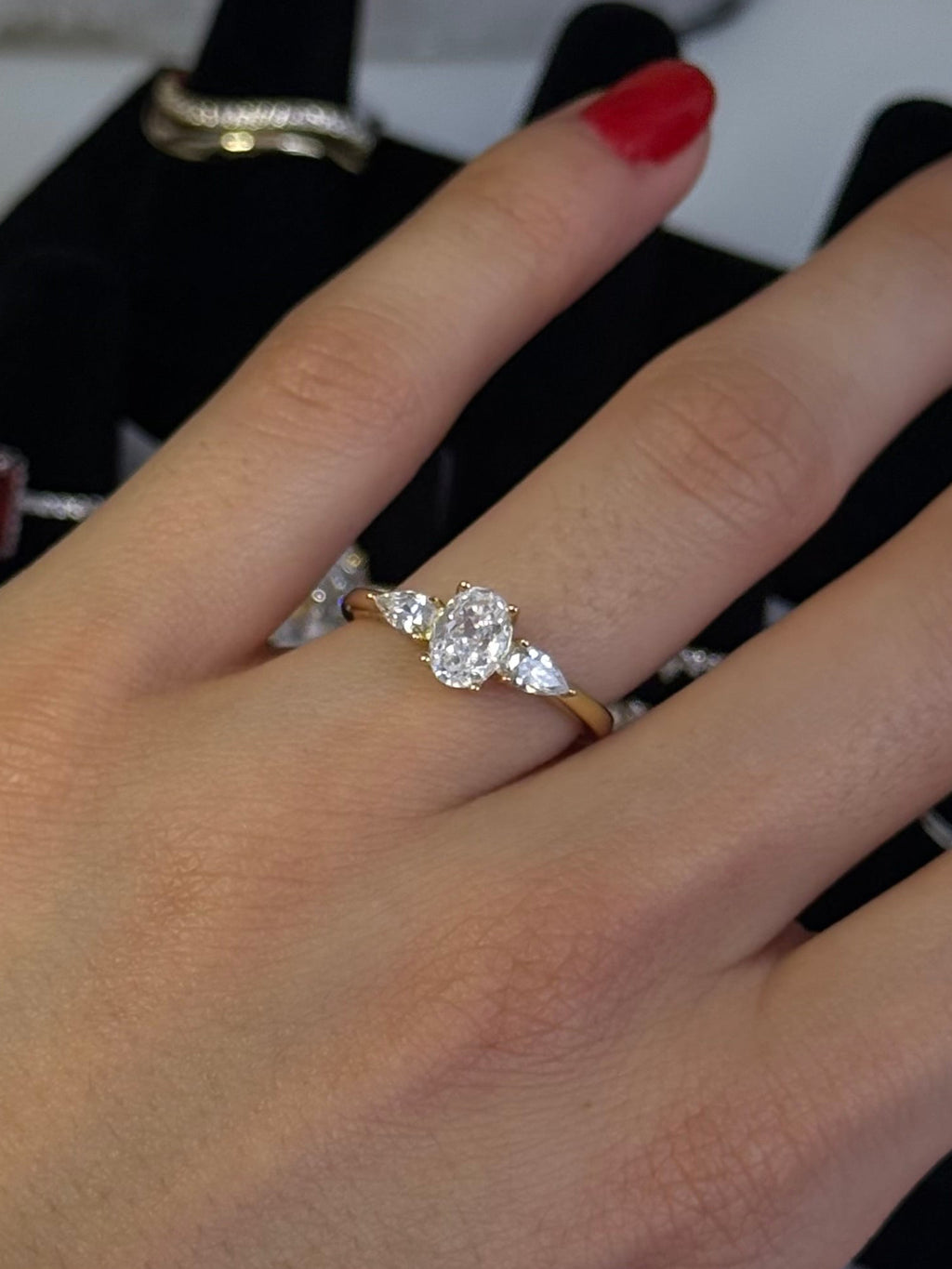 A close-up of a hand wearing a silver ring with a central round-cut diamond and smaller side stones, along with gold Roman numeral bangles. The hand is resting on a black jewelry display stand with other rings in the background.