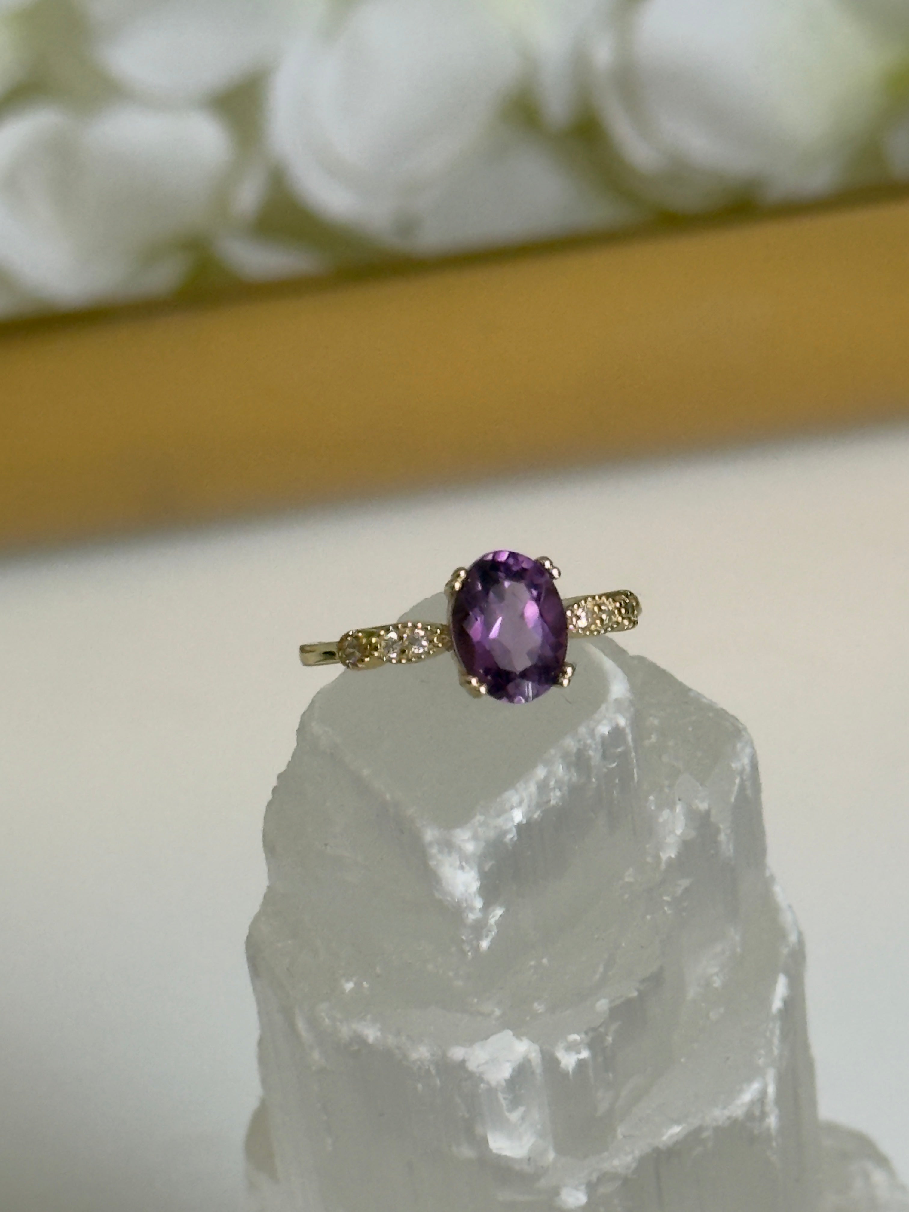 Gold ring with a purple gemstone on a clear crystal with white flowers in the background