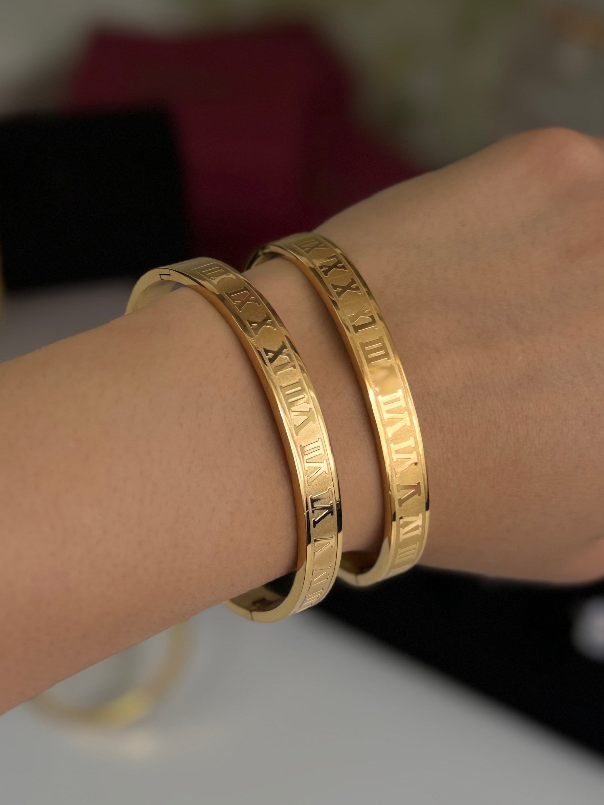 Two gold bangle bracelets with engraved Roman numerals worn on a person's wrist, shown against a softly blurred background.